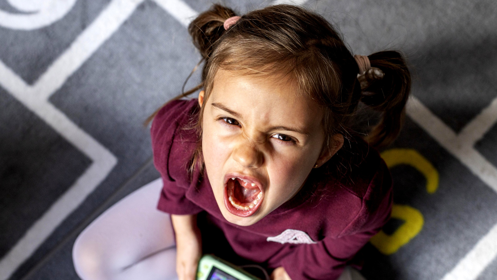 Bambina seduta su di un tappeto che guarda all'insù e urla.