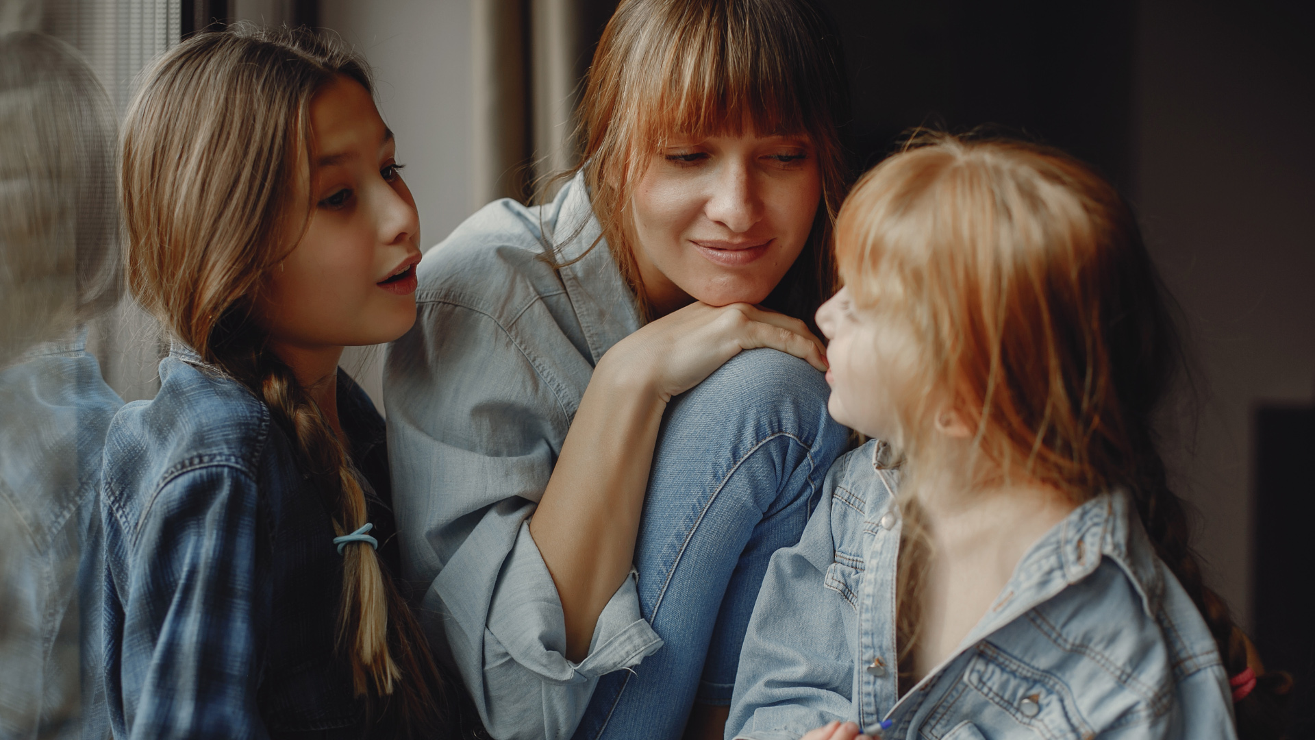 Mamma accovacciata con due figlie a conversare con volto dolce e interessato.